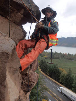 Ben George inspecting a rock slope via rope access high above I-84.
